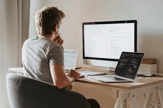 student sitting at a desk in front of three computer monitors, working on multiple programming assignments simultaneously